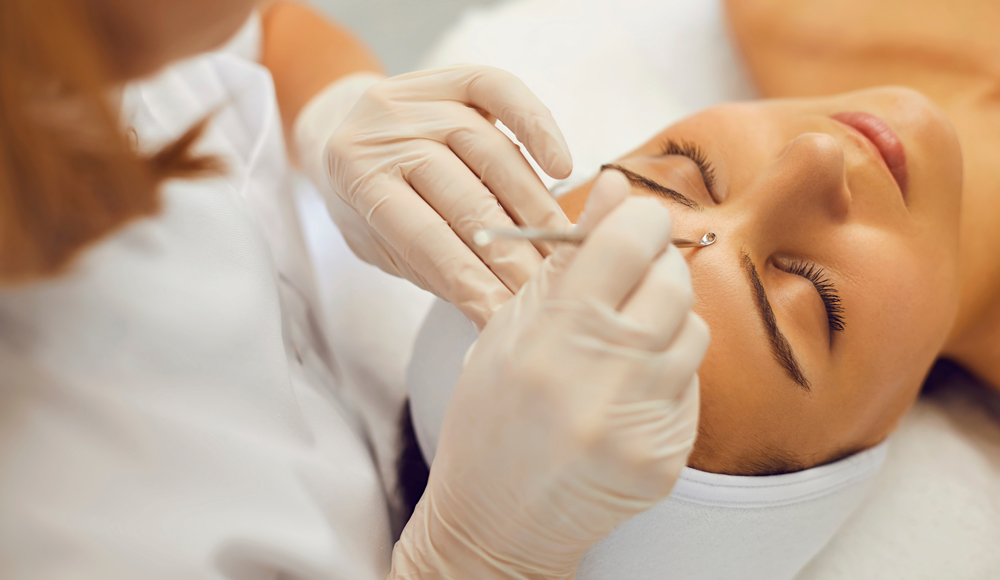 High angle of cosmetician using blackhead and whitehead remover to make skin of female client clear and healthy. Beautician in medical gloves removing pimples on face of customer with steel extractor