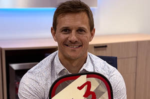 Smiling man in a gray patterned shirt holding a pickleball paddle